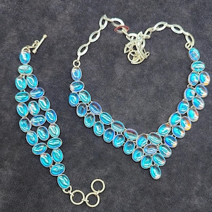 May include: A silver-toned necklace and bracelet set. Both pieces feature iridescent, oval-shaped blue gemstones. The gemstones have a shimmering, multi-coloured effect, reflecting hues of blue and orange. The necklace and bracelet are connected by silver-toned metal.