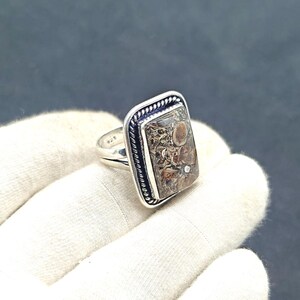 May include: A silver ring with a rectangular, patterned stone. The stone displays brown, tan, and cream hues, with a distinctive, natural pattern. The ring has a textured border and a plain band.
