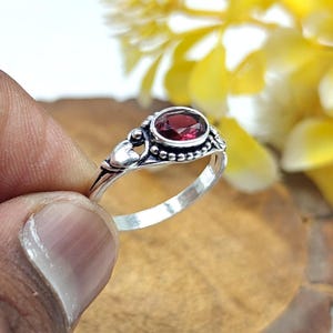 May include: A silver ring featuring an oval, deep red gemstone. The ring has a detailed design with small heart-shaped accents and beaded edges. The gemstone is set in a bezel setting, and the ring is held by a person's fingers.