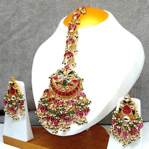 以下が含まれることがあります： 金色のインドのジュエリーセット。ヘッドピースとイヤリングが含まれています。ヘッドピースは、赤、緑、白のアクセントと、ぶら下がっている赤と白のビーズを備えた中央の円形デザインです。イヤリングは、同様の色とビーズ細工でデザインを反映しています。