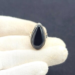 Op de afbeelding: Zilveren ring met een grote, zwarte, druppelvormige edelsteen. De gepolijste steen is gezet in een gedetailleerde zilveren band met decoratieve randen. De ring wordt getoond tegen een witte achtergrond.
