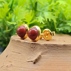 Puede incluir: Un par de pendientes de botón dorados con una piedra redonda de color rojo intenso engastada en un bisel dorado. Los pendientes se muestran sobre una superficie marrón con un fondo verde borroso.