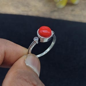 May include: A silver ring featuring a prominent, smooth, round, bright red stone set in a raised bezel. The band includes a small, clear, circular accent stone on one side, positioned near the main setting, with a polished metallic finish.