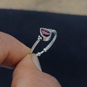 May include: A silver ring featuring a teardrop-shaped dark red gemstone set in a metal bezel. The band has a polished finish with small decorative metal spheres on either side of the stone setting and additional bead-like accents along the shank.