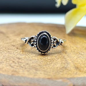 Op de afbeelding: Een zilveren ring met een ovale, zwarte edelsteen. De ring heeft een sierlijk ontwerp rond de edelsteen en op de band. De ring ligt op een houten oppervlak. Sieraden.