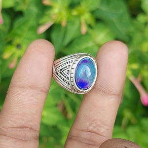Puede incluir: Un anillo de metal plateado con una piedra de humor ovalada que muestra un degradado de colores azul y morado vibrantes. La banda está decorada con intrincados patrones triangulares y de puntos grabados en la superficie metálica alrededor.