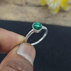 Könnte beinhalten: Ein Silberring mit einem kleinen, runden, polierten Malachit-Edelstein in einer einfachen Zargenfassung. Der grüne Stein zeigt natürliche dunkle Bänderungen, und die Ringschiene besteht aus glattem, poliertem Silber mit einem dünnen Profil.