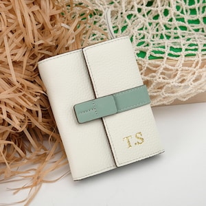 May include: A white leather wallet with a mint green strap closure. The wallet has the gold initials "T.S" embossed on the front. The wallet is closed and has a textured surface. The background includes brown paper and a white net bag.