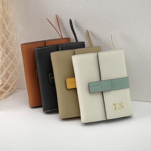 May include: A row of four leather wallets in various colors: brown, black, tan, and white. Each wallet has a strap closure and a loop for a wristlet. The white wallet has the initials "T.S" in gold lettering.