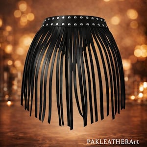 Puede incluir: Falda de cuero negro con cinturilla tachonada y flecos largos. La falda tiene forma redondeada y está hecha de tiras de cuero. El texto "PAKLEATHERArt" es visible en la parte inferior. El fondo está borroso con luces bokeh.