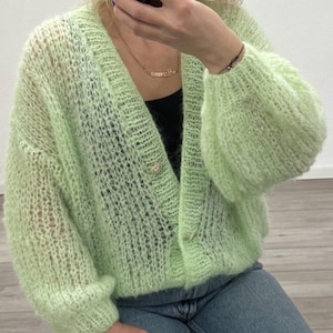 Könnte beinhalten: Hellgrüne Strickjacke mit offenem Webmuster, V-Ausschnitt und langen, puffärmeligen Ärmeln. Die Strickjacke hat zwei goldfarbene Knöpfe und wird mit einer blauen Jeans kombiniert. Die Strickjacke ist ein weicher, flauschiger Strick.