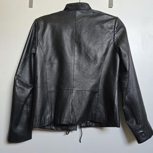 May include: A black leather jacket with a mandarin collar and long sleeves. The back of the jacket is visible, showing the seams and a drawstring detail at the waist. The jacket is hanging on a white background.