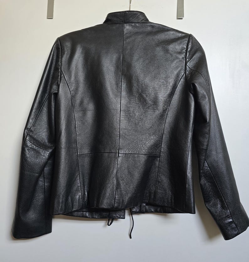 May include: A black leather jacket with a mandarin collar and long sleeves. The back of the jacket is visible, showing the seams and a drawstring detail at the waist. The jacket is hanging on a white background.