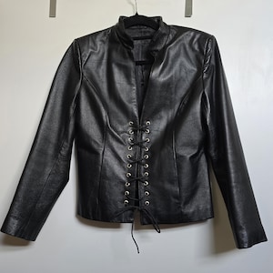 May include: Black leather jacket with a stand-up collar and long sleeves. The front features a lace-up closure with gold-colored eyelets. The jacket is hanging on a black hanger against a white background. A stylish outerwear piece.