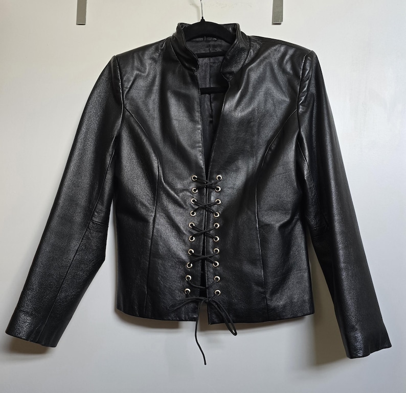 May include: Black leather jacket with a stand-up collar and long sleeves. The front features a lace-up closure with gold-colored eyelets. The jacket is hanging on a black hanger against a white background. A stylish outerwear piece.
