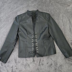 May include: Black leather jacket with a stand-up collar and long sleeves. The front features a lace-up closure with silver-toned eyelets. The jacket is laid flat on a gray surface.
