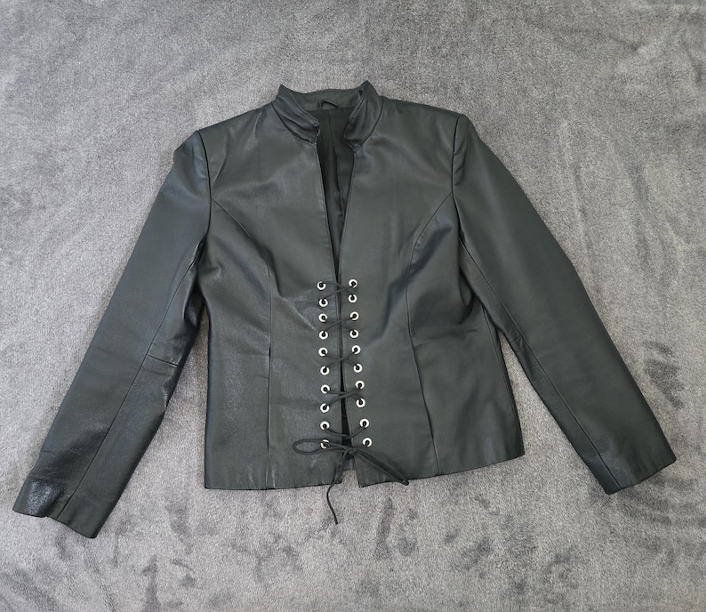 May include: Black leather jacket with a stand-up collar and long sleeves. The front features a lace-up closure with silver-toned eyelets. The jacket is laid flat on a gray surface.