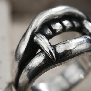 May include: A silver-toned ring featuring a sculpted design of a mouth with prominent fangs. The metal has a polished finish with darkened recessed areas to highlight the detailed shape of the teeth and the curved structure of the band.
