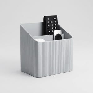 May include: A pale grey, ribbed desk tidy with a sloping top. It contains a black remote control, a white remote, and a small white item. The organiser is designed to hold various items, keeping them neat and tidy.