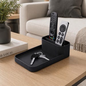 May include: A black, ribbed-textured organiser on a wooden table. It holds two remote controls and a set of keys. The organiser has a tray section and a divided compartment for remotes. A plant in a dark vase and a book are also on the table.