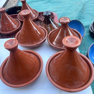May include: A collection of terracotta tagine pots, featuring conical lids and rounded bases. The pots are a warm reddish-brown color, with a few in the background showing a darker brown hue. Some blue and black bowls are also visible.
