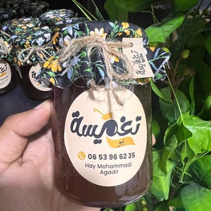 Puede incluir: Un tarro de miel con una tapa de tela floral negra atada con hilo. El tarro tiene una etiqueta blanca con texto árabe y un número de teléfono. La miel es de color ámbar oscuro. Otros tarros son visibles en el fondo.