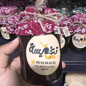 Puede incluir: Frascos de vidrio llenos de un líquido oscuro, cada uno sellado con una tapa de tela burdeos atada con hilo. Los frascos tienen una etiqueta redonda con texto árabe e información de contacto.