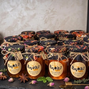 May include: A collection of small glass jars filled with golden honey. Each jar is adorned with a black floral fabric lid, tied with twine, and a label with Arabic text. The jars are arranged on a dark surface, with dried flowers and a honey dipper.