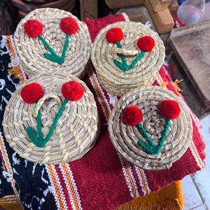May include: Four round woven coasters with a cherry blossom design. Each coaster features a woven straw base, red pom-pom cherries, and green embroidered stems and leaves. The coasters rest on a red and black patterned textile.