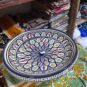 May include: A round, ceramic plate with a detailed, colorful floral design. The plate features a dark blue rim and a central pattern with red, yellow, and green accents. The plate is sitting on a green and yellow textile.