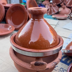May include: A brown ceramic tagine pot with a conical lid and a wide base. The pot has a shiny glaze and a silver band around the middle. Other similar pots and plates are visible in the background.