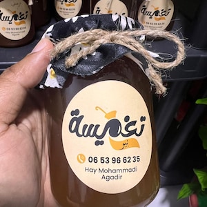 Puede incluir: Un tarro de miel con una tapa de tela floral en blanco y negro atada con hilo. El tarro tiene una etiqueta redonda con texto árabe y un número de teléfono. La miel es de color marrón dorado.