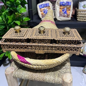 Puede incluir: Una caja decorativa de madera con tres compartimentos, cada uno coronado con un pomo dorado. La caja se encuentra sobre una cesta tejida y un pequeño taburete. Detrás hay bolsas de comida y un sombrero tejido.