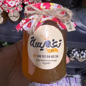 Puede incluir: Un tarro de miel con una tapa de tela estampada con flores atada con hilo de bramante. El tarro tiene una etiqueta con texto árabe y un número de teléfono. La miel es de color ámbar dorado.