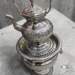 以下が含まれることがあります： 装飾的なデザインの銀色のティーポット。 ティーポットには注ぎ口、ハンドル、蓋が付いています。 装飾的な模様のある段になったベースの上にあります。 全体的なデザインは華やかで詳細です。 高さ約25cm。