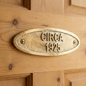 May include: A brass oval plaque with the text "CIRCA 1925" is mounted on a light-colored wooden door. The plaque is secured with two screws, and the wood grain is visible.