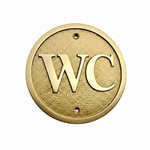 Könnte beinhalten: Rundes, goldfarbenes Metallschild mit den Buchstaben "WC" in fetter Schrift. Das Schild hat einen strukturierten Hintergrund und zwei kleine Löcher zur Befestigung. Es ist ein Toilettenschild.