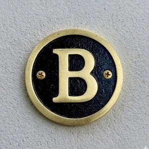 以下が含まれることがあります： 金色の文字「B」が入った真鍮製の丸い表札。文字は黒い背景の中央に配置され、金色の縁で囲まれています。2本の真鍮ネジで固定されています。