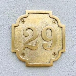 May include: A brass house number plaque with the number 29. The plaque is rectangular with decorative edges and is secured with four screws. The numbers are in a classic serif font.