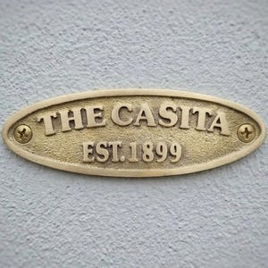 Könnte beinhalten: Goldfarbene ovale Plakette mit den geprägten Worten "THE CASITA" und "EST. 1899". Die Plakette ist mit zwei Schrauben an einer hellgrauen, strukturierten Wand befestigt. Der Text ist fett und in einer Serifenschrift.