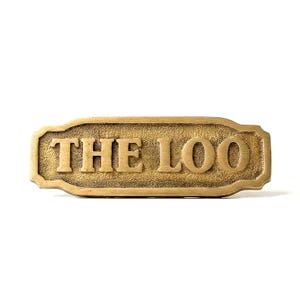 May include: A rectangular, antique brass sign with the text "THE LOO" in bold, raised letters. The sign has a decorative border and a textured surface, suggesting an aged appearance. Ideal for bathroom decor.