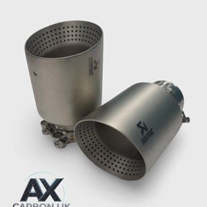 May include: Two silver exhaust tips with the Akrapovic logo and multiple small holes. One tip is upright, and the other is lying on its side. The text "AX CARBON UK" is at the bottom.