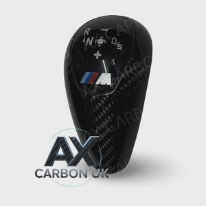 May include: A black carbon fibre gear shift knob with the BMW M logo. The knob has the letters R, N, D/S, and a plus sign. The logo "AX CARBON UK" is at the bottom.