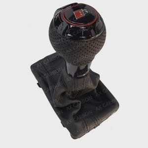 May include: A black and red car gear shift knob with a perforated grip. The top features a red ring and a logo. The base is covered in black leather. The text "AX CARBON" is visible on the leather.