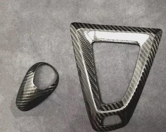May include: Two carbon fiber car interior trim pieces. One is a triangular frame with a white border, and the other is a small, oval-shaped piece. Both have a black and gray woven pattern.