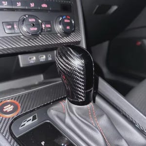 May include: A carbon fibre and black gear shift knob in a car interior. The shift knob has a glossy finish and is surrounded by a grey leather boot with red stitching. The dashboard features carbon fibre trim and various controls.