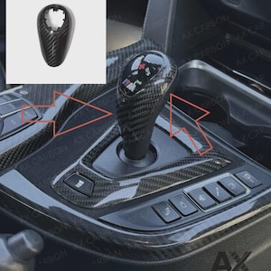 May include: Black carbon fibre gear shift knob and console trim. The gear shift knob has an M logo. The console has buttons for various car functions. The trim has a carbon fibre weave pattern.