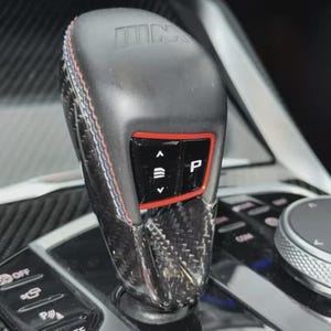 May include: A close-up of a car gear shift lever. The lever is black with carbon fibre accents and red stitching. The top of the lever has the letters "M" and "W" printed on it. The lever has a red-bordered control panel with buttons.