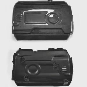 May include: Two black engine covers. The top cover has a carbon fibre weave pattern, a circular indentation, and rectangular cutouts. The bottom cover is solid black with a circular indentation and a series of vertical slits.