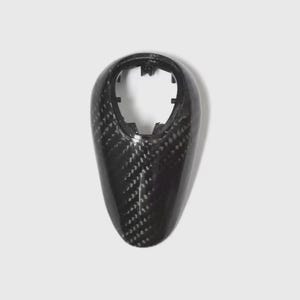 May include: A carbon fibre gear shift knob with a glossy finish. The knob has a teardrop shape with a hollowed-out top for attachment. The carbon fibre weave is visible, creating a textured appearance.